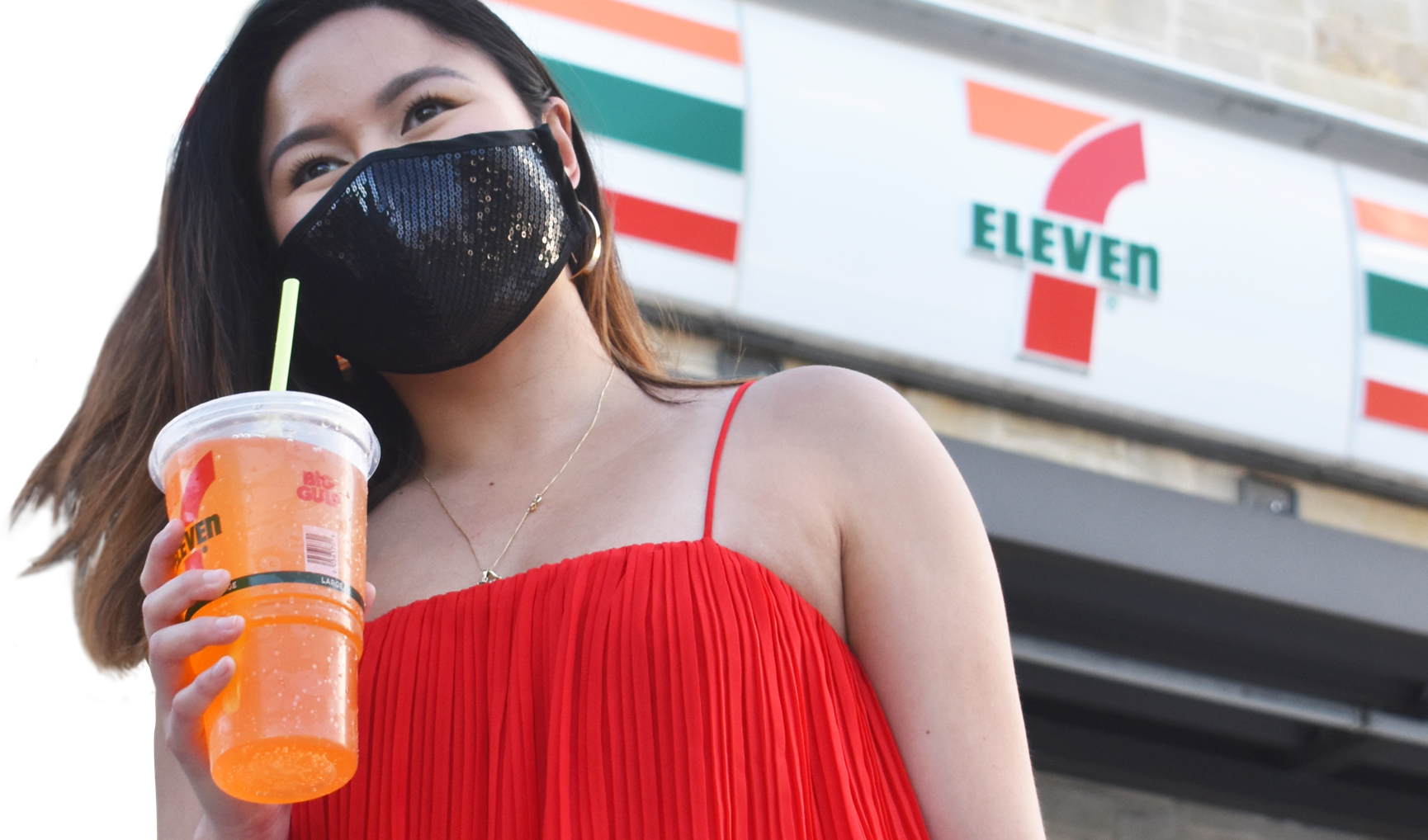 A young woman with a black sequin face mask and red top, holding an orange colored Big Gulp, outside a 7-Eleven store.